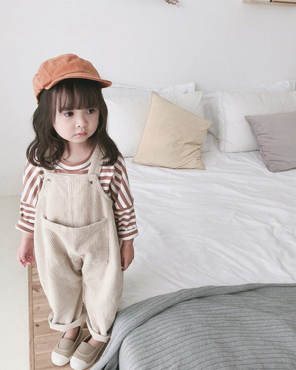 Petite fille portant une superbe salopette ample en velours côtelé fille de couleurs beige avec une marinière, des baskets et une casquette terracotta dans une chambre, devant un lit.