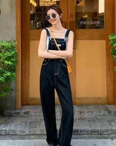 on voit à l'extérieur devant une porte d'entrée en bois une jeune femme brune portant une salopette en jean