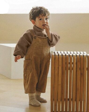 Photo d'un petit garçon portant une salopette en velours côtelé marron, il se tient devant un cube en bois avec une main à la bouche