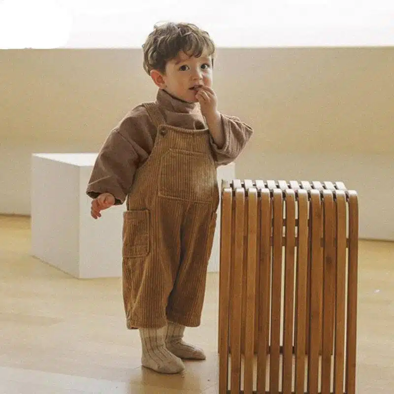 Photo d'un petit garçon portant une salopette en velours côtelé marron, il se tient devant un cube en bois avec une main à la bouche