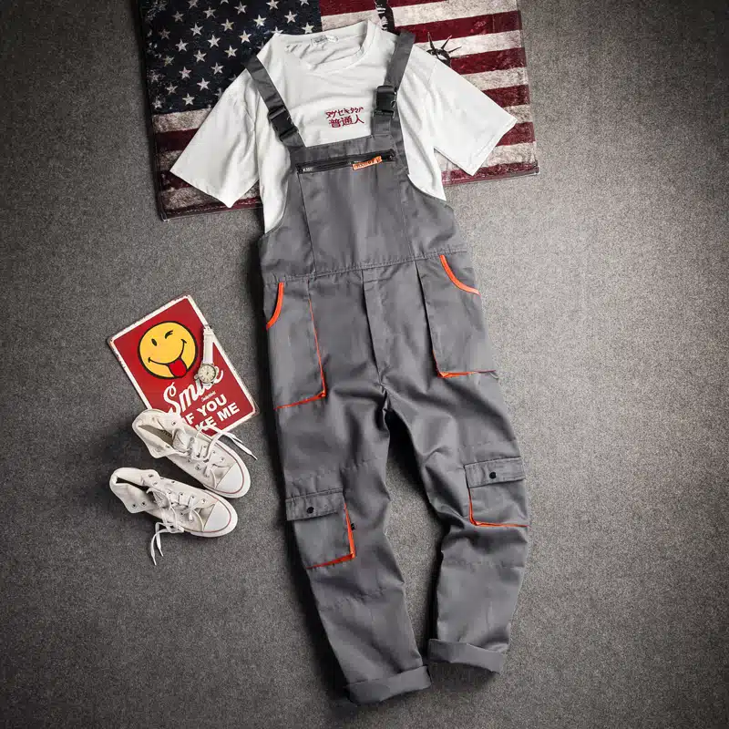 Sur fond gris foncé, près d'une paire de baskets blanches et sur un drapeau américain, on voit une salopette de travail grise avec un liseré orange au niveau des poches.