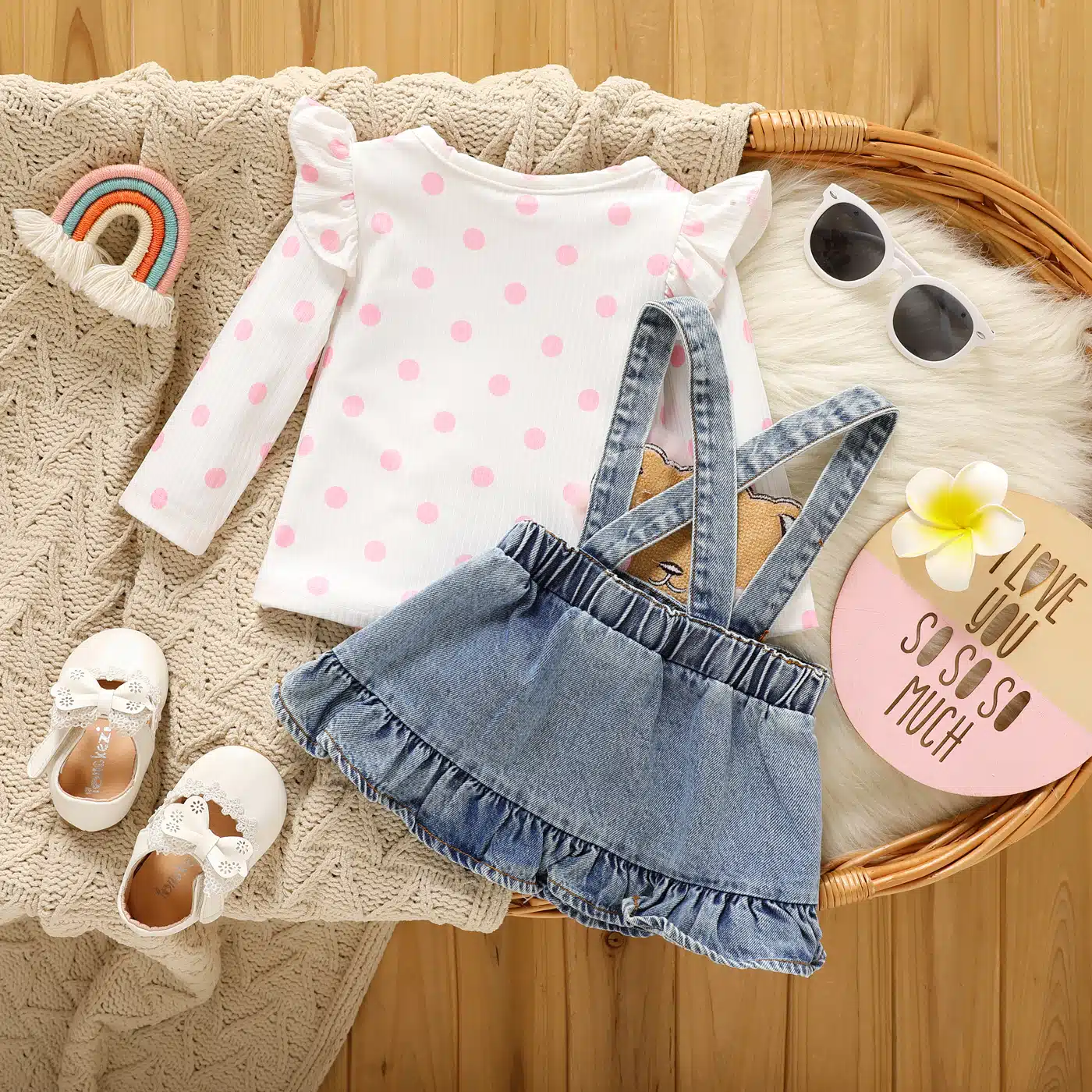 Sur un meuble, on voit une corbeille à langer dans laquelle sont joliment exposés un plaid beige et une tenue composée d'une tee-shirt manche longue blanc et rose et d'une jupe-salopette pour bébé.