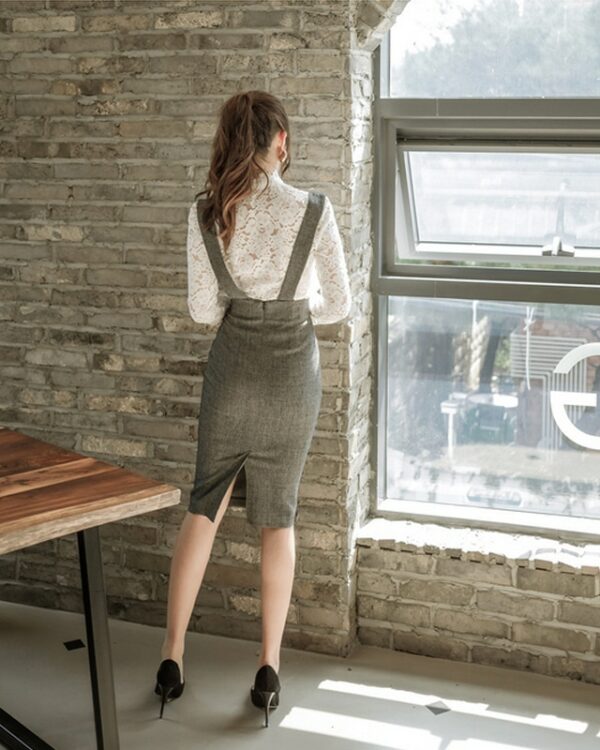 On voit une jeune femme brune aux cheveux longs, de dos, qui regarde par une baie vitrée et qui porte une jupe-salopette assortie à un chemisier en dentelle blanc. Le look fait très business woman.