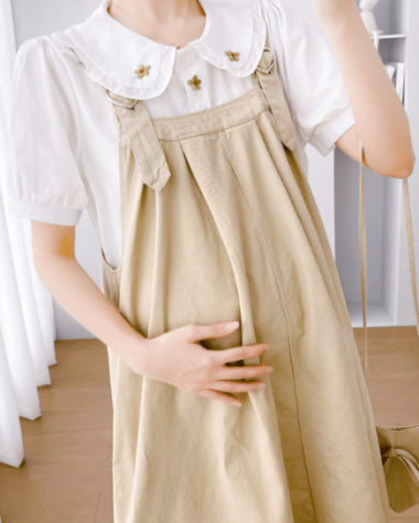 On voit une femme qui porte un chemisier avec une robe salopette beige. Elle est enceinte et porte sa main à son ventre.
