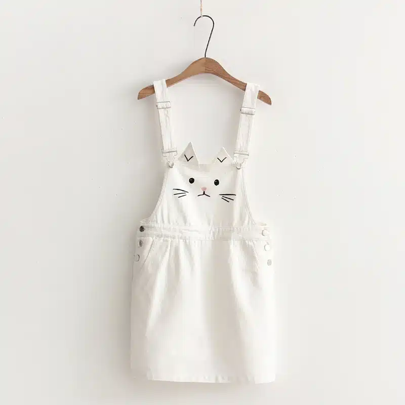 Sur fond blanc, on voit une jupe-salopette blanche avec un motif tête de chat. Elle est suspendue à un cintre en bois.
