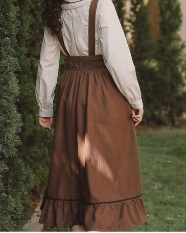 On voit une jeune femme de dos au look très romantique. Elle porte une chemise blanche avec une jupe salopette marron, son look est très vintage. Elle marche dans un jardin.
