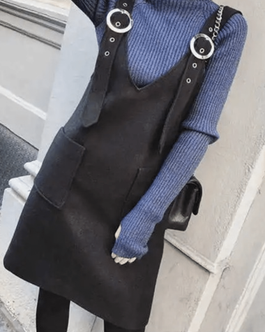 Photo d'une robe salopette droite avec un col en V et des bretelles façon ceintures, la robe salopette est noir portée par une femme avec un sous-pull violet, des collants noir et un sac à main à chaine.