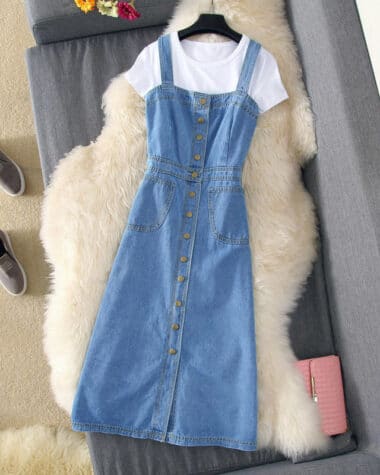 Robe salopette en jean, cintré à la taille, boutonnière avant, portée sur un cintre avec un tee-shirt blanc. La robe est posée sur une fourrure blanche sur un canapé gris.