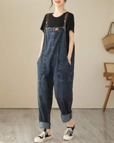 Photo d'une femme portant une salopette large en jean avec des bretelles marron, des baskets noires et un t-shirt noir. La femme se tient debout avec les mains dans les poches, dans une pièce aux teintes beige avec une chaise en rotin et un panier en arrière plan.