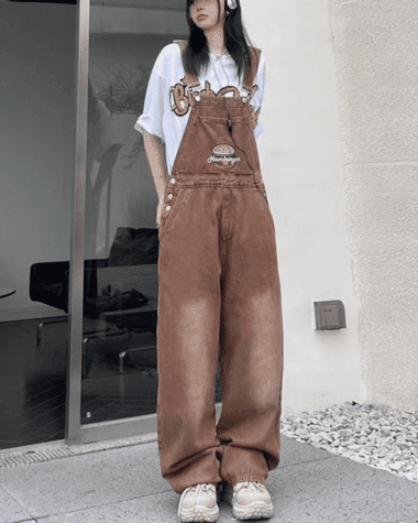 Photo d'une femme portant une salopette en jean marron avec un hamburger brodé sur le torse, elle se tient debout devant un immeuble.