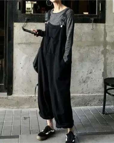 Femme portant une salopette large noire en jean avec un haut ris et des vans, elle se tien dans la rue devant un café