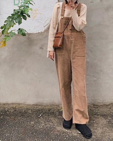 Photo d'une femme portant une salopette large en velours côtelé beige et un look nude. Elle se tient debout devant un mur avec des feuilles d'arbres.