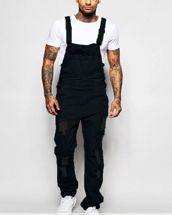Homme tatoué portant une salopette noire en jean déchiré sur les jambes avec un t-shirt et des baskets blancs