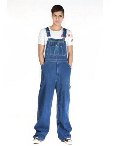 Salopette jean homme confortable bleu avec étiquette verte sur la poche centrale portée par un jeune homme vêtue d'un t-shirt blanc et de sneakers.